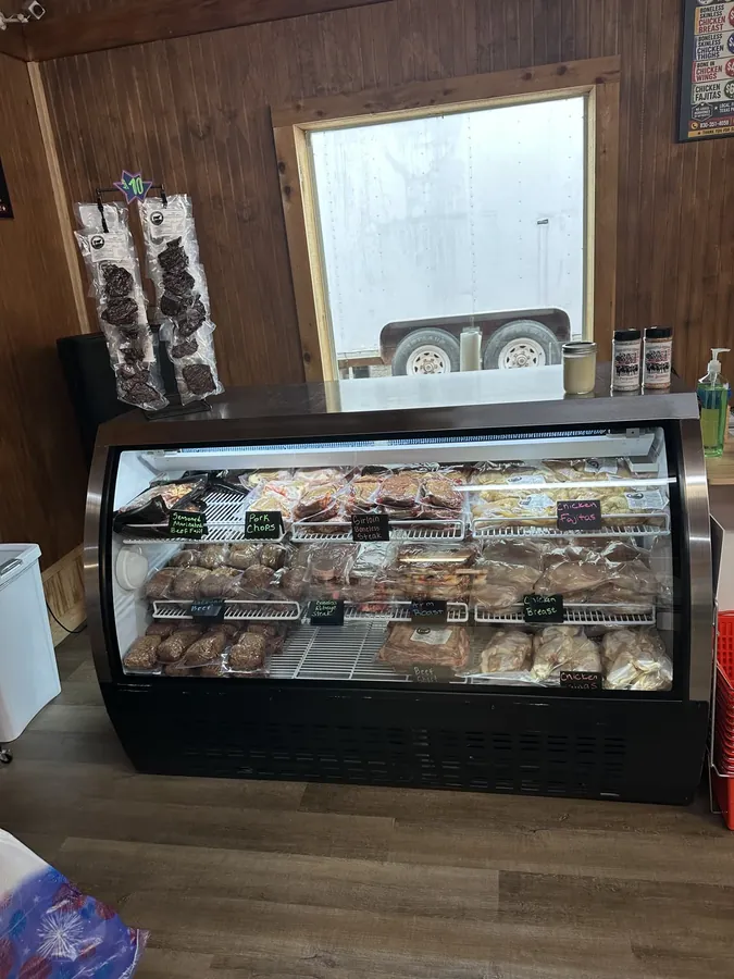 The refrigerated case stocked with hand-labeled cuts including ground beef, boneless ribeye steak, arm roast and chicken breast, with racks of vacuum-sealed beef jerky and jars of 3-M seasoning on top.