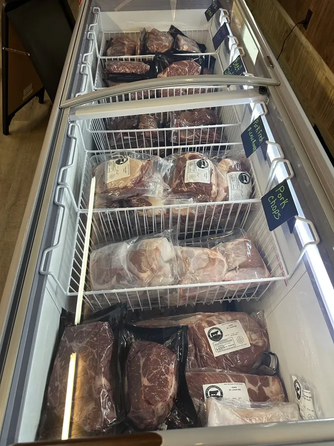 Open chest freezer with wire baskets of vacuum-sealed pork chops, fresh picnic roast, St. Louis ribs and beef cuts, each basket marked with a chalkboard tag.