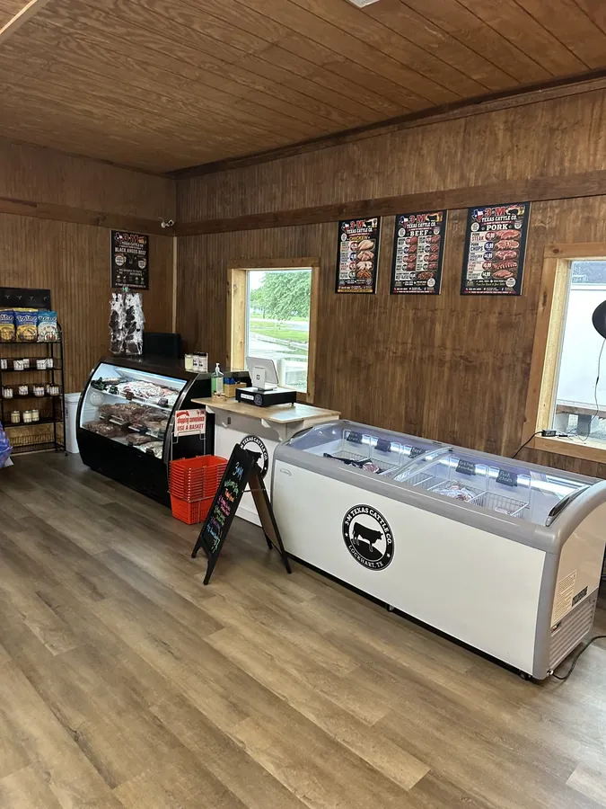 Inside the 3-M Texas Cattle Co. store in Lockhart: a refrigerated deli case of vacuum-sealed cuts on the left, a chest freezer carrying the 3-M bull logo on the right, and beef, pork, and chicken price boards on the back wall.